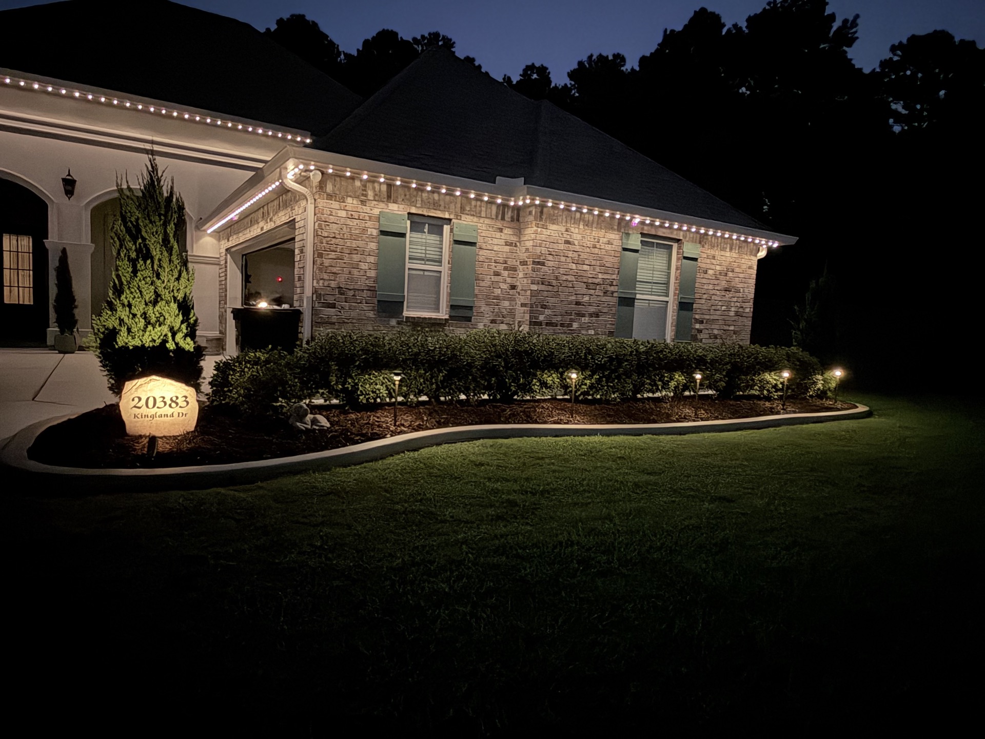 Brick clubhouse at dusk with warm roofline lighting and a row of path lights illuminating a curved landscape bed and address marker