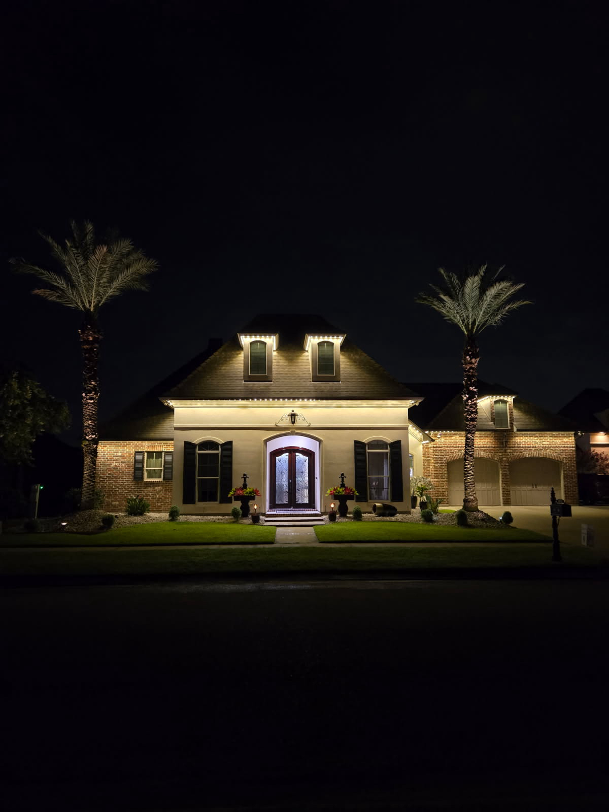 Symmetrical estate-style building at night with dormer roofline lighting, uplit palm trees, and a glowing entry door
